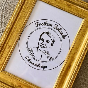 May include: A gold-colored picture frame with a white mat and a black and white logo inside. The logo features a woman's face and the text "Froilein Jolanda Schmuckdesign".