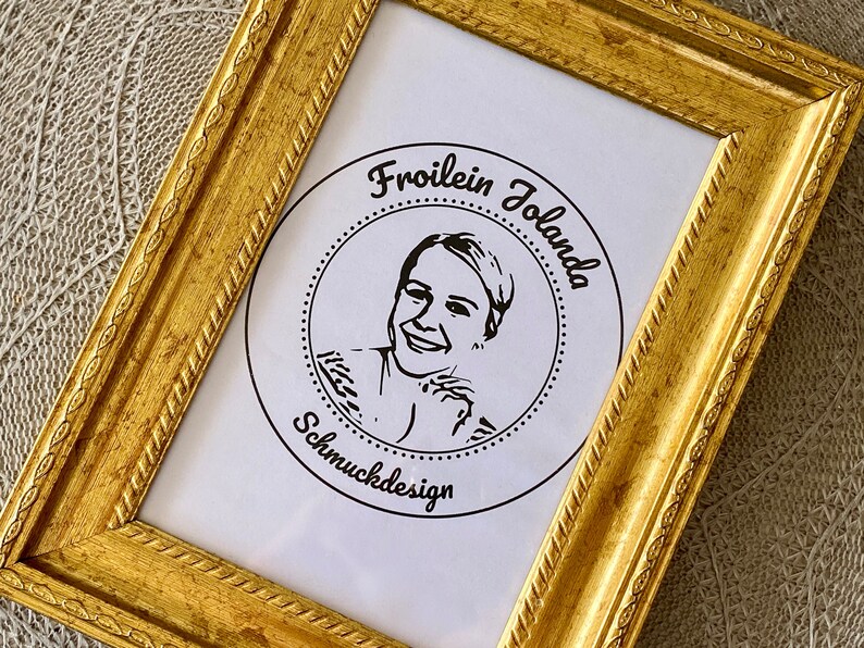 May include: A gold-colored picture frame with a white mat and a black and white logo inside. The logo features a woman's face and the text "Froilein Jolanda Schmuckdesign".