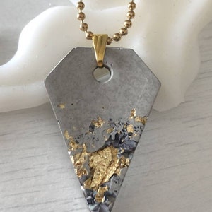 May include: A gray concrete pendant with gold flecks, attached to a gold chain. The pendant is shaped like a diamond.