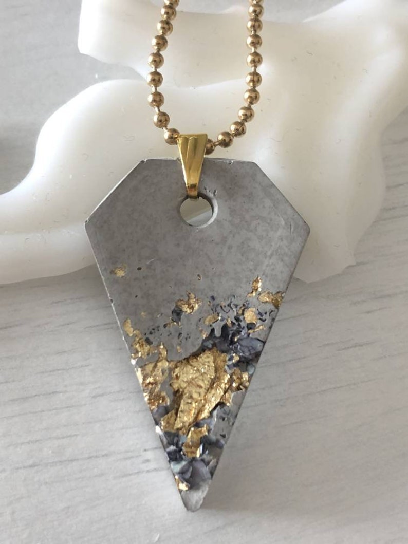 May include: A gray concrete pendant with gold flecks, attached to a gold chain. The pendant is shaped like a diamond.