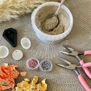 May include: A variety of craft supplies for making jewelry, including a container of gray powder, two small containers of colorful beads, two silicone molds, a round piece of white material, a pair of pink pliers, and a pair of silver pliers.