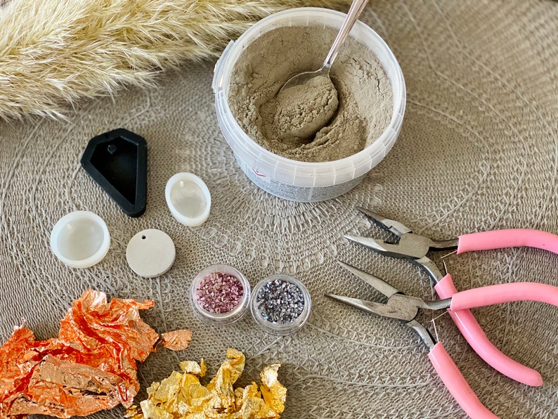 May include: A variety of craft supplies for making jewelry, including a container of gray powder, two small containers of colorful beads, two silicone molds, a round piece of white material, a pair of pink pliers, and a pair of silver pliers.