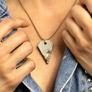May include: A gold chain necklace with a gray concrete pendant featuring gold flecks. The pendant is shaped like a triangle with rounded edges.