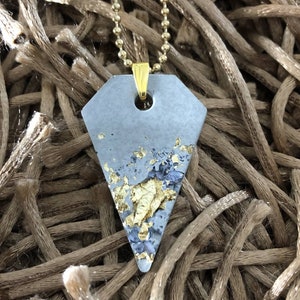 May include: A gray concrete pendant with gold leaf accents hangs from a gold chain. The pendant is shaped like a diamond and has a hole in the top for the chain to pass through.