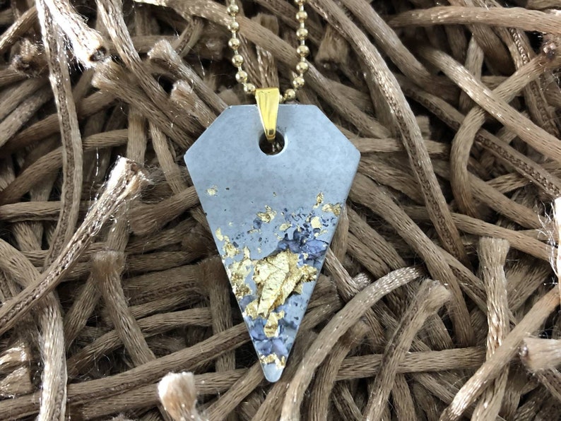 May include: A gray concrete pendant with gold leaf accents hangs from a gold chain. The pendant is shaped like a diamond and has a hole in the top for the chain to pass through.