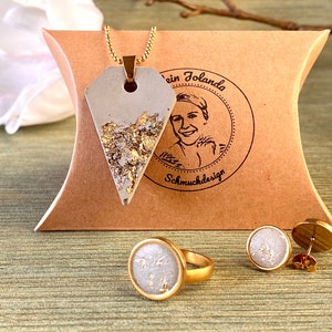 Könnte beinhalten: Ein Set aus Goldschmuck mit einem Betonanhänger mit Blattgold-Akzenten, einem Ring mit einer runden Betonplatte und einem Paar Ohrsteckern. Der Schmuck ist in einer braunen Schachtel mit einem Logo, das "ein Yolanda Schmuckdesign" lautet, ausgestellt.