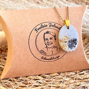 May include: A gold chain necklace with a light gray oval pendant featuring gold and black flecks. The pendant is in a small brown box with a logo that reads "Froilein Joland Schmuckdesign".