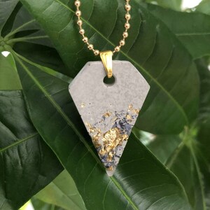 May include: A gray concrete pendant with gold flecks on a gold chain. The pendant is shaped like a triangle with rounded edges.