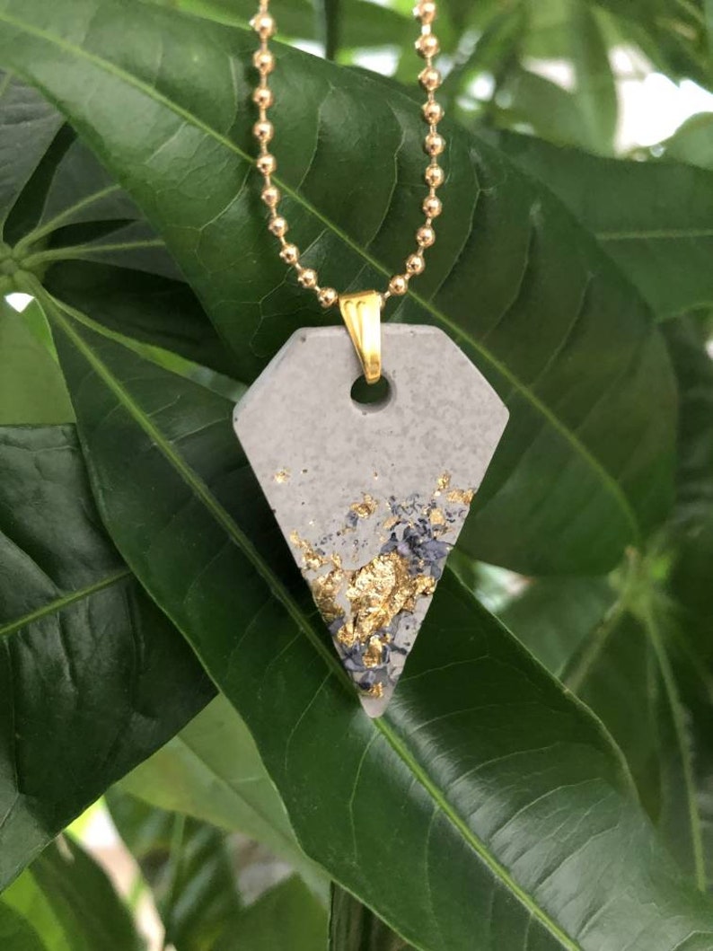 May include: A gray concrete pendant with gold flecks on a gold chain. The pendant is shaped like a triangle with rounded edges.