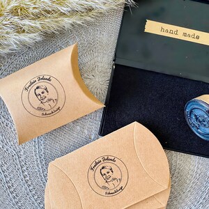 May include: Two brown paper envelopes with a black and white circular stamp that says "Fraulein Jolando Schmuckdesign" and a black and white image of a woman. The envelopes are on a gray surface. A black ink pad with a wooden stamp is also on the surface. The ink pad has a label that says "hand made".