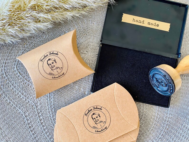 May include: Two brown paper envelopes with a black and white circular stamp that says "Fraulein Jolando Schmuckdesign" and a black and white image of a woman. The envelopes are on a gray surface. A black ink pad with a wooden stamp is also on the surface. The ink pad has a label that says "hand made".