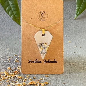 May include: A gold chain necklace with a white triangular pendant featuring gold glitter. The pendant is on a brown card with the text "Fräulein Jolanda" printed on it.