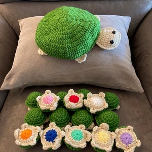 May include: A collection of 12 crocheted turtle toys. The turtles are green with a variety of colored shells. The turtles are arranged in a circle on a brown surface.