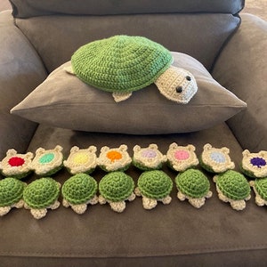 May include: A collection of 14 crocheted turtle toys, each with a different colored shell. The turtles are arranged in a row on a gray couch. There is a larger crocheted turtle toy in the background.