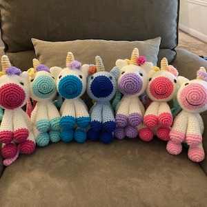 May include: Seven crocheted unicorn toys in various colors, including pink, blue, purple, and yellow. The unicorns are sitting on a light brown couch.