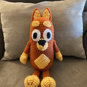 May include: A crocheted stuffed animal in the shape of a dog. The dog is brown with yellow accents and black eyes. It is sitting on a gray couch.