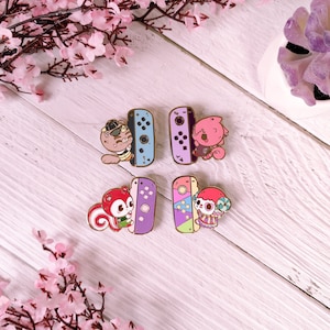 May include: Four enamel pins featuring characters from the Animal Crossing video game series. Each pin features a different character holding a Nintendo Switch console. The characters are a squirrel, a cat, a dog, and a sheep. The pins are colorful and have a glossy finish.