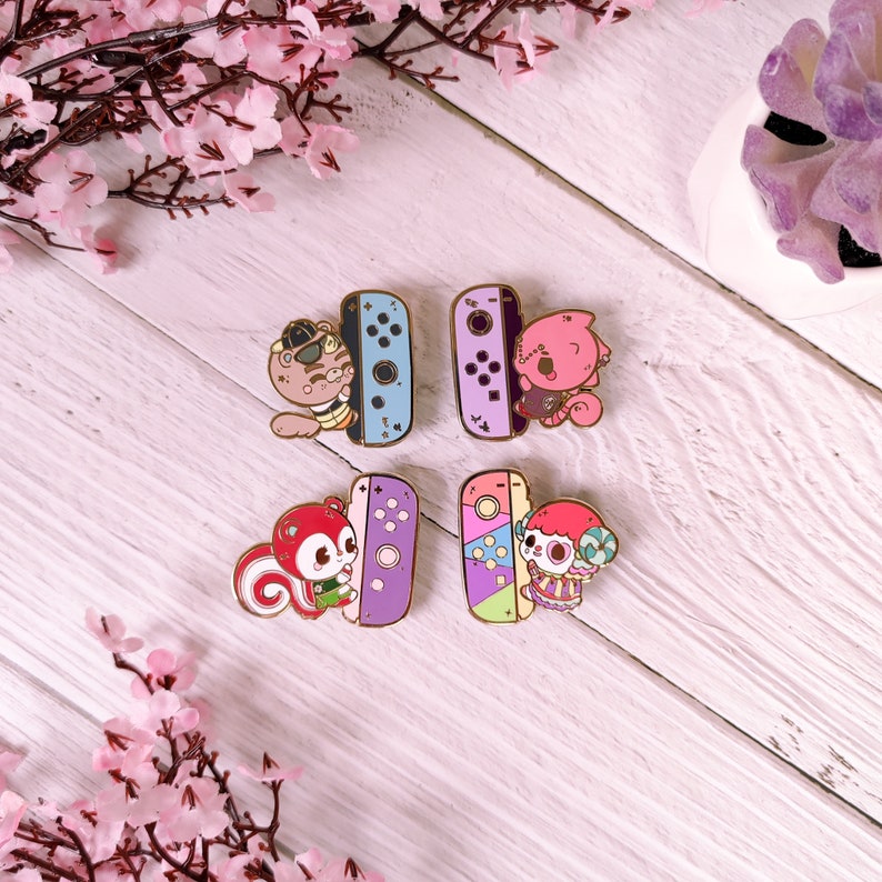 May include: Four enamel pins featuring characters from the Animal Crossing video game series. Each pin features a different character holding a Nintendo Switch console. The characters are a squirrel, a cat, a dog, and a sheep. The pins are colorful and have a glossy finish.
