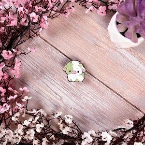 May include: A green and white enamel pin featuring a cartoon cow with a pink nose and black eyes. The pin is on a light brown wooden surface surrounded by pink and white flowers.