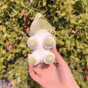 May include: A small plush toy in the shape of a cat with green and white fur. The toy has a bell attached to its neck.