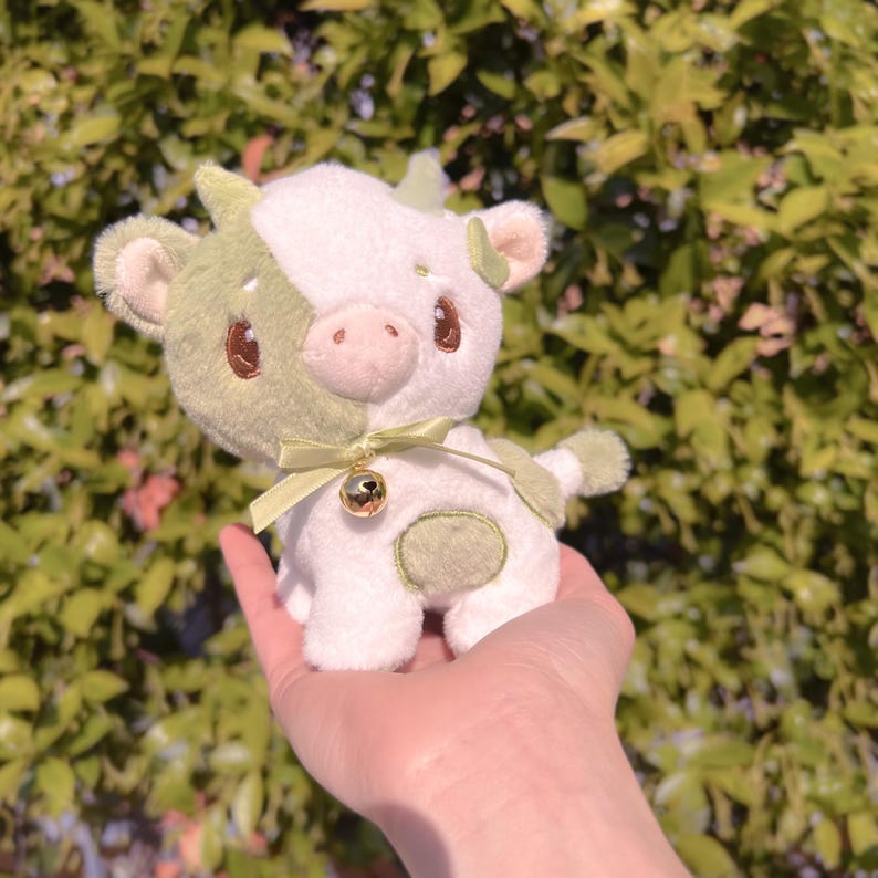 May include: A plush green and white cow toy with a bell around its neck. The toy has a cute, cartoonish face and is held in a person's hand.