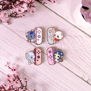 May include: Four enamel pins featuring cartoon animal characters holding a pink and blue Nintendo Switch controller. The characters are a blue cat, a white cat, a blue and white cat, and a blue and white cat with a purple and pink dress.