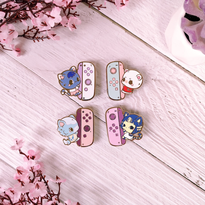 May include: Four enamel pins featuring cartoon animal characters holding a pink and blue Nintendo Switch controller. The characters are a blue cat, a white cat, a blue and white cat, and a blue and white cat with a purple and pink dress.