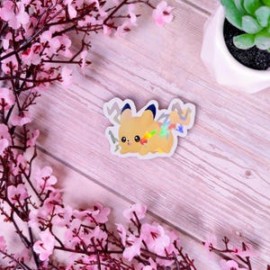 Puede incluir: Una pegatina holográfica con un personaje de dibujos animados con pelaje amarillo, orejas con puntas azules y una cola de rayo. La pegatina tiene un diseño de arcoíris. Fondo de madera rosa claro, con flores rosas y una pequeña planta suculenta.