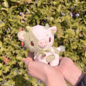 May include: A plush toy cow with a white body and green spots. The cow has a bell around its neck and is being held in someone's hands.