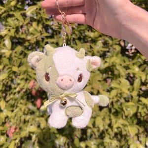 May include: A plush green and white cow keychain with a gold bell. The cow has a cute face with big eyes and a pink nose.