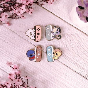 May include: Four enamel pins featuring animal characters from the Animal Crossing video game series. Each pin depicts a different character holding a Nintendo Switch Joy-Con controller. The characters are a white dog, a black cat, a blue bird, and a brown squirrel.
