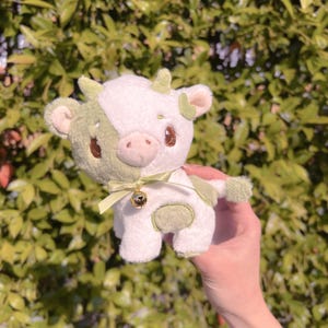May include: A plush toy cow with a white and green patchwork design. The cow has a green bow and a gold bell around its neck.