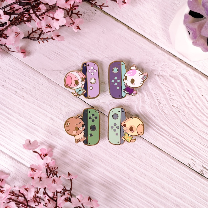 May include: Four enamel pins featuring characters from the Animal Crossing video game series. Each pin depicts a character holding a Nintendo Switch console. The characters are a pink bunny, a brown bear, a white dog, and a white cat. The consoles are in different colors: purple, green, and blue.