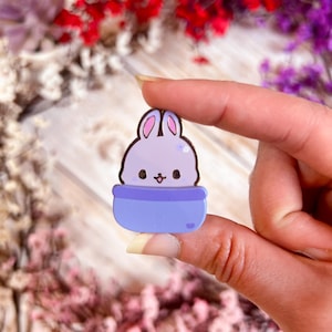 May include: A cute, lavender bunny in a blue pot-shaped design. The bunny has pink inner ears and a small, smiling face. The pot has a heart detail. This enamel pin is a charming accessory.