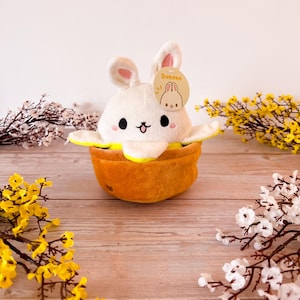 May include: A white plush bunny wearing a yellow banana peel costume. The bunny has black eyes, a pink nose, and a smiling mouth. The banana peel is brown and has a yellow lining. The bunny is sitting on a wooden surface with yellow and white flowers in the background.