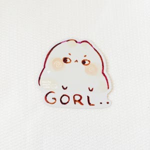 May include: A white sticker featuring a cartoon character with a rounded shape, rosy cheeks, and small eyes. The sticker has the text "GORI.." in brown letters. The sticker also has the text "mocha mochi cake" in a smaller font.