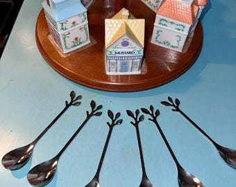 May include: A collection of ceramic house-shaped spice jars with various colored roofs, arranged on a wooden carousel. The jars are decorated with floral patterns. Below, six stainless steel spoons with leaf-shaped handles are displayed.