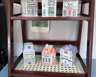 May include: A wooden shelf unit displays six ceramic spice jars shaped like miniature houses. The jars are white with colorful details, including text labels like "Mustard" and "Relish." The shelf has a dark brown finish.
