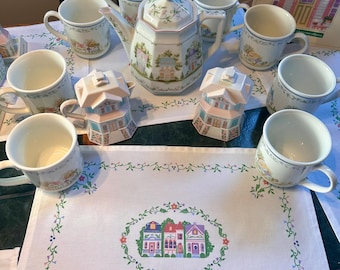 Op de afbeelding: Een Lenox Village theeservies met een theepot, mokken en huisvormige room- en suikerpotten. De witte porseleinen stukken zijn versierd met kleurrijke huis motieven en bloemaccenten. De set wordt gepresenteerd op een wit tafelkleed met een bijpassend geborduurd ontwerp.