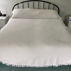 May include: A white bedspread with a textured waffle weave and a fringe border. The bedspread is on a black metal bed frame with a curved headboard.