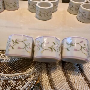 K&ouml;nnte beinhalten: Wei&szlig;e Serviettenringe aus Keramik mit einem floralen Design aus gr&uuml;nen Bl&auml;ttern und rosa Bl&uuml;ten. Die Serviettenringe haben einen hellvioletten Rand und sind auf einer dekorativen Oberfl&auml;che angeordnet. Ein Set eleganter Tischdekoration.