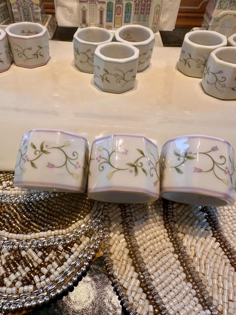 K&ouml;nnte beinhalten: Wei&szlig;e Serviettenringe aus Keramik mit einem floralen Design aus gr&uuml;nen Bl&auml;ttern und rosa Bl&uuml;ten. Die Serviettenringe haben einen hellvioletten Rand und sind auf einer dekorativen Oberfl&auml;che angeordnet. Ein Set eleganter Tischdekoration.