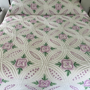 May include: A white cotton bedspread with a raised floral pattern. The flowers are a light purple with green leaves. The bedspread has a scalloped edge.