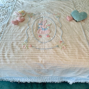 May include: A white and blue crocheted throw blanket with a pink embroidered girl holding flowers in a blue oval frame. The blanket has a blue and white striped pattern and a fringe border. There are three pink hearts and a blue heart on the blanket.