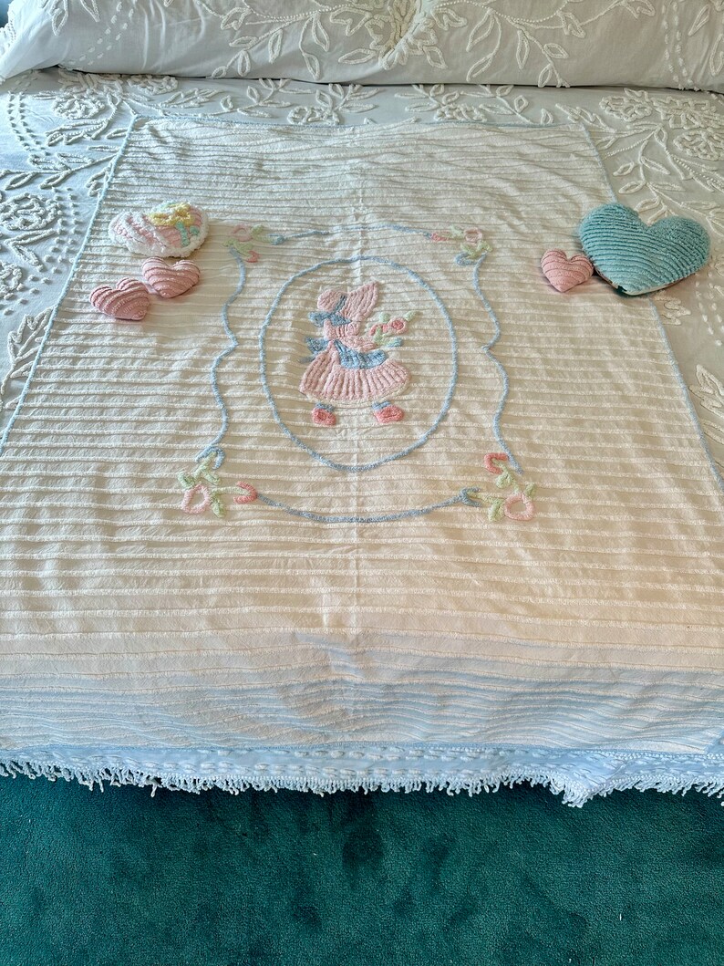 May include: A white and blue crocheted throw blanket with a pink embroidered girl holding flowers in a blue oval frame. The blanket has a blue and white striped pattern and a fringe border. There are three pink hearts and a blue heart on the blanket.