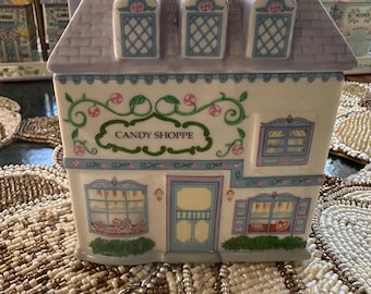 Op de afbeelding: Een keramische Candy Shoppe-container, in de vorm van een huis met een grijs dak en dakkapellen. De gevel is wit met blauwe en roze accenten, en de woorden "CANDY SHOPPE" staan op de voorkant.