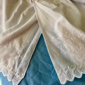 May include: A light cream-colored garment featuring an elasticized gathered neckline and a scalloped hem. The fabric is adorned with intricate white floral embroidery and eyelet cutouts that follow a delicate vine pattern along the edges.