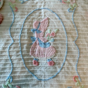 May include: A white crocheted blanket with a blue and pink embroidered design of a girl holding flowers. The blanket has a striped pattern and is decorated with floral accents.
