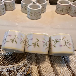 K&ouml;nnte beinhalten: Eine Sammlung wei&szlig;er Keramik-Serviettenringe mit floralem Design. Die Ringe haben einen gelben Rand und sind mit gr&uuml;nen Bl&auml;ttern und lila Blumen verziert. Die Serviettenringe sind auf einer wei&szlig;en Oberfl&auml;che angeordnet.