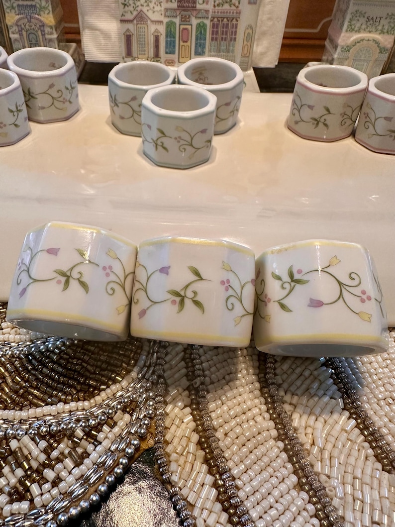 K&ouml;nnte beinhalten: Eine Sammlung wei&szlig;er Keramik-Serviettenringe mit floralem Design. Die Ringe haben einen gelben Rand und sind mit gr&uuml;nen Bl&auml;ttern und lila Blumen verziert. Die Serviettenringe sind auf einer wei&szlig;en Oberfl&auml;che angeordnet.